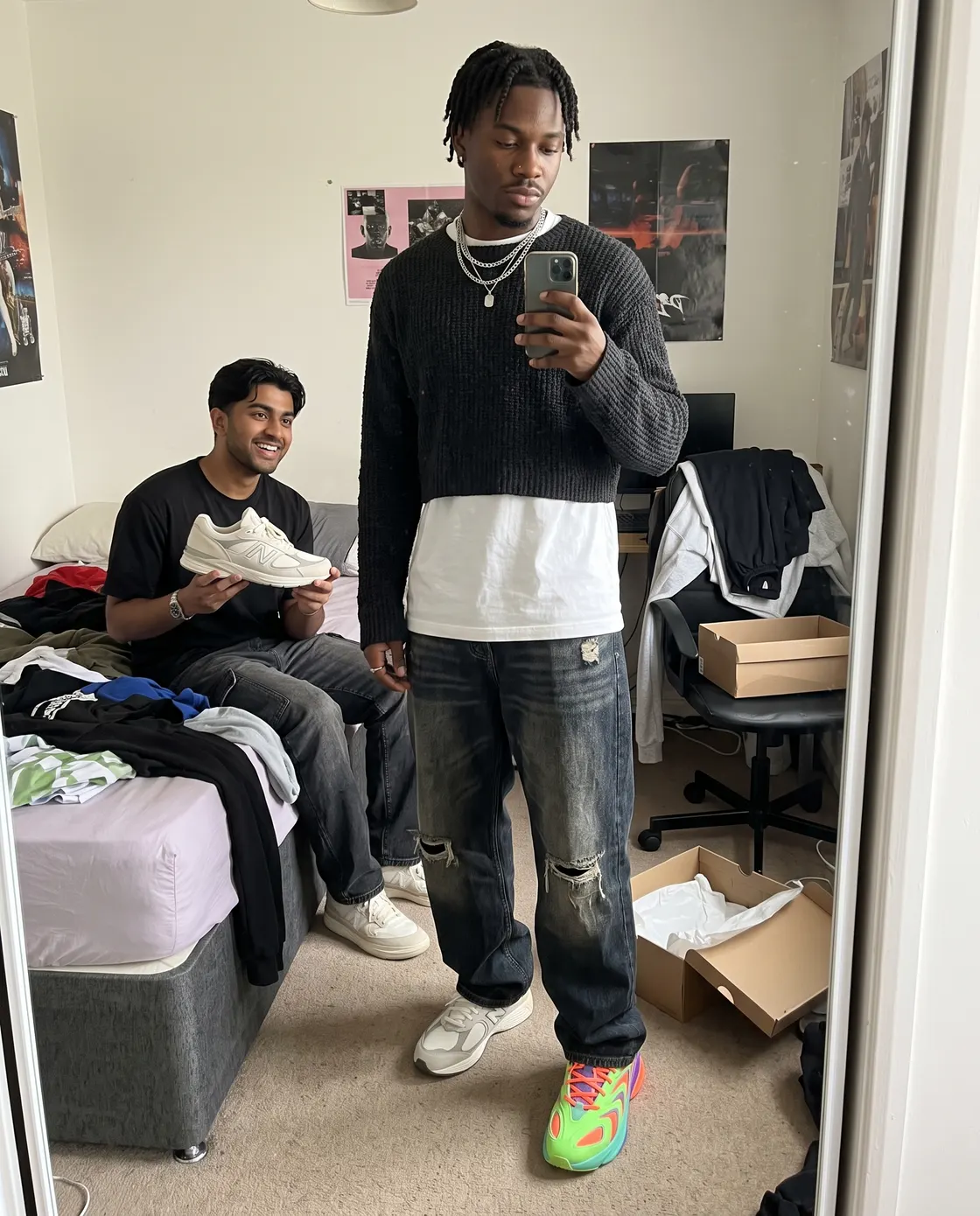 A man taking a bedroom mirror photo wearing a different trainer on each foot while a friend holds up a third option.