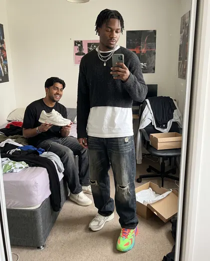 A man taking a bedroom mirror photo wearing a different trainer on each foot while a friend holds up a third option.