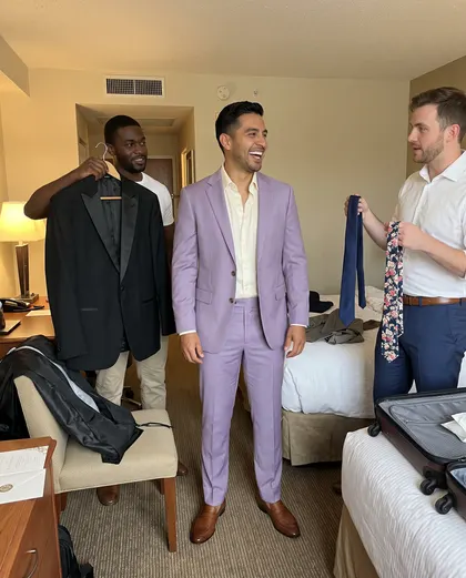 A man in a lilac suit in a hotel room while one friend holds up a black tuxedo jacket and another offers two ties.