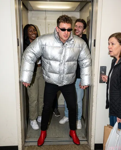 A man in an enormous silver puffer jacket and red cowboy boots filling a lift doorway while two friends laugh behind him and a neighbour stares.